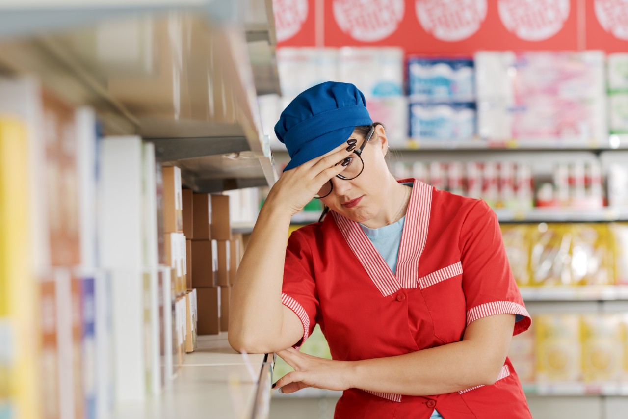 Tired exhausted supermarket employee leaning on a store shelf and resting, she is having a headache