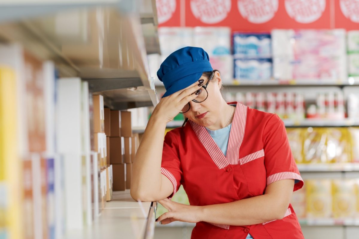 Tired exhausted supermarket employee leaning on a store shelf and resting, she is having a headache