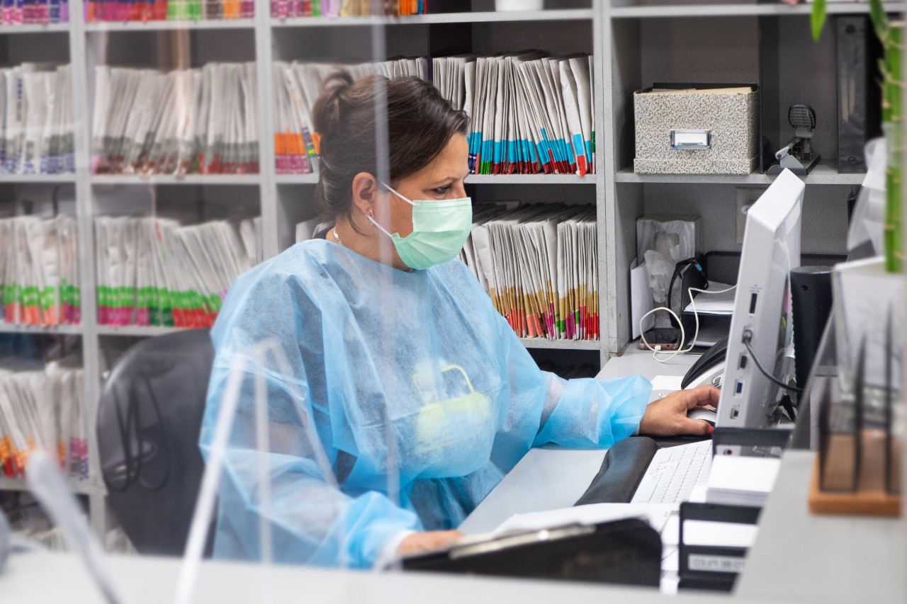 Hispanic receptionist at dental office wearing mask and gown for protection during the Covid-19 pandemic. She is working at the computer with patient files in the background.