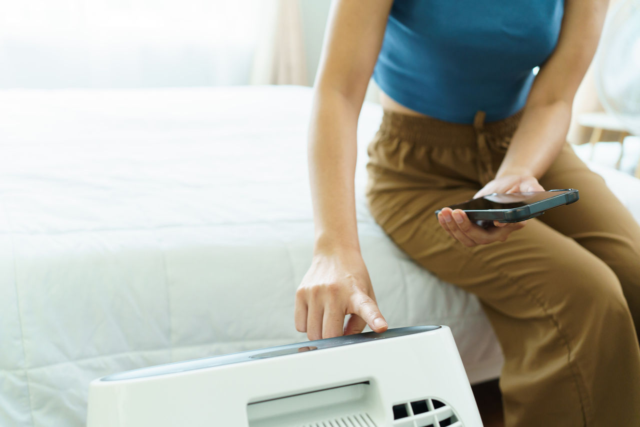 woman sitting on the bed in bedroom and using air purifier, woman using application on a smartphone to control a digital wireless air purifier machine.