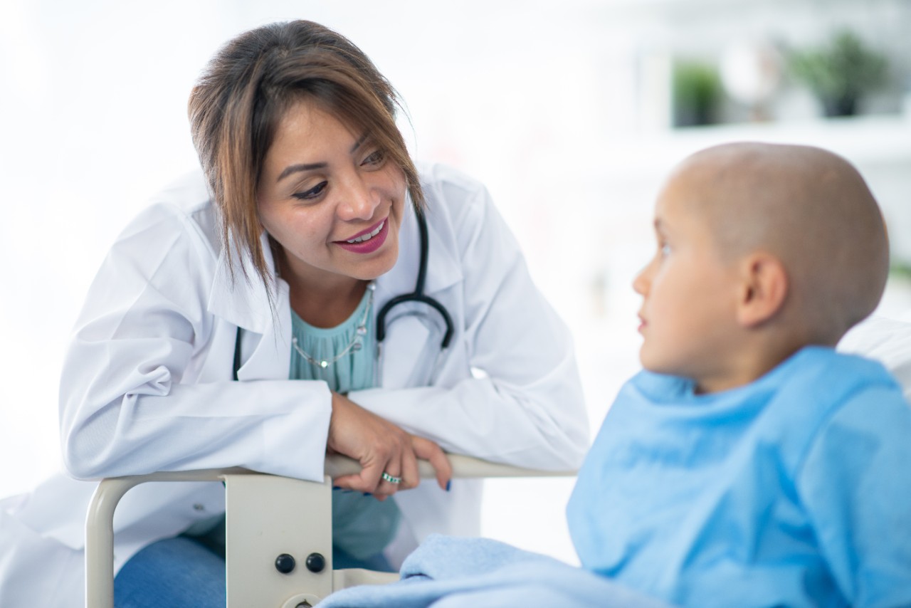 A young boy with cancer sits up in his hospital bed, wearing a blue gown and with a downcast expression on his face.  His female Hispanic doctor leans in over the bed rail, dressed in a white lab coat, as she talks with him.