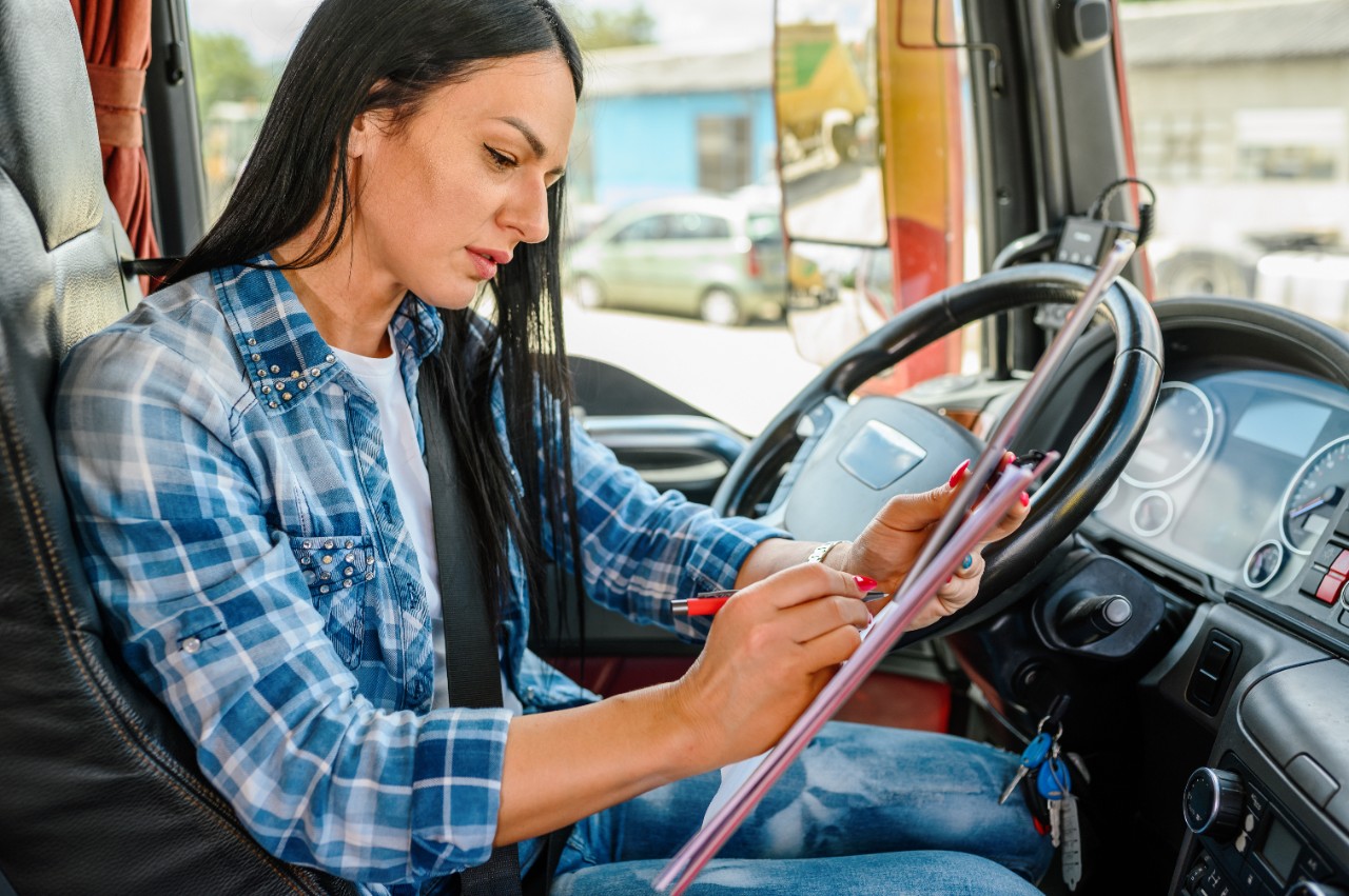 A professional female truck driver who using a digital tablet and writing on a clipboard in the cab of a truck